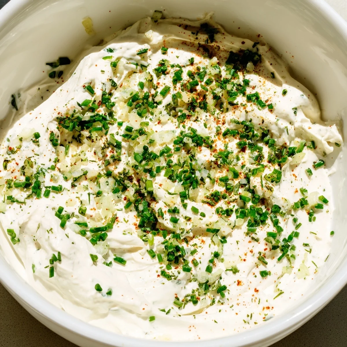 A bowl of creamy dip garnished with fresh chives and paprika, served with sliced cucumbers, carrots, and pita chips.