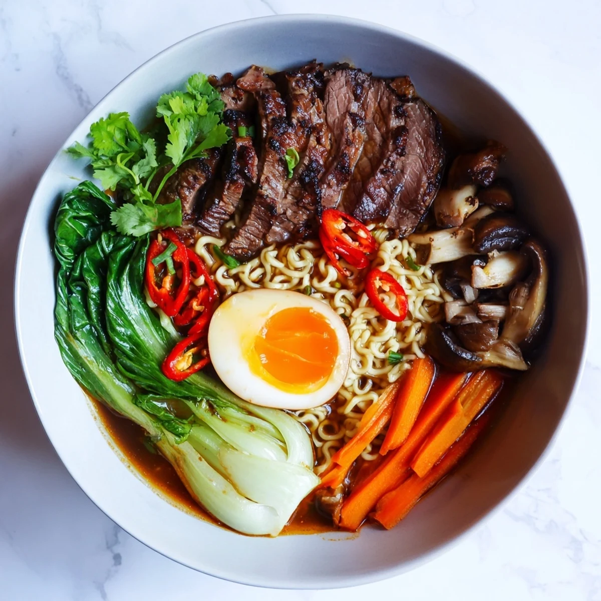 Overhead view of Spicy Beef Ramen with Soft Boiled Egg, showcasing julienned carrots, bok choy, and a halved jammy egg next to a steaming bowl.
