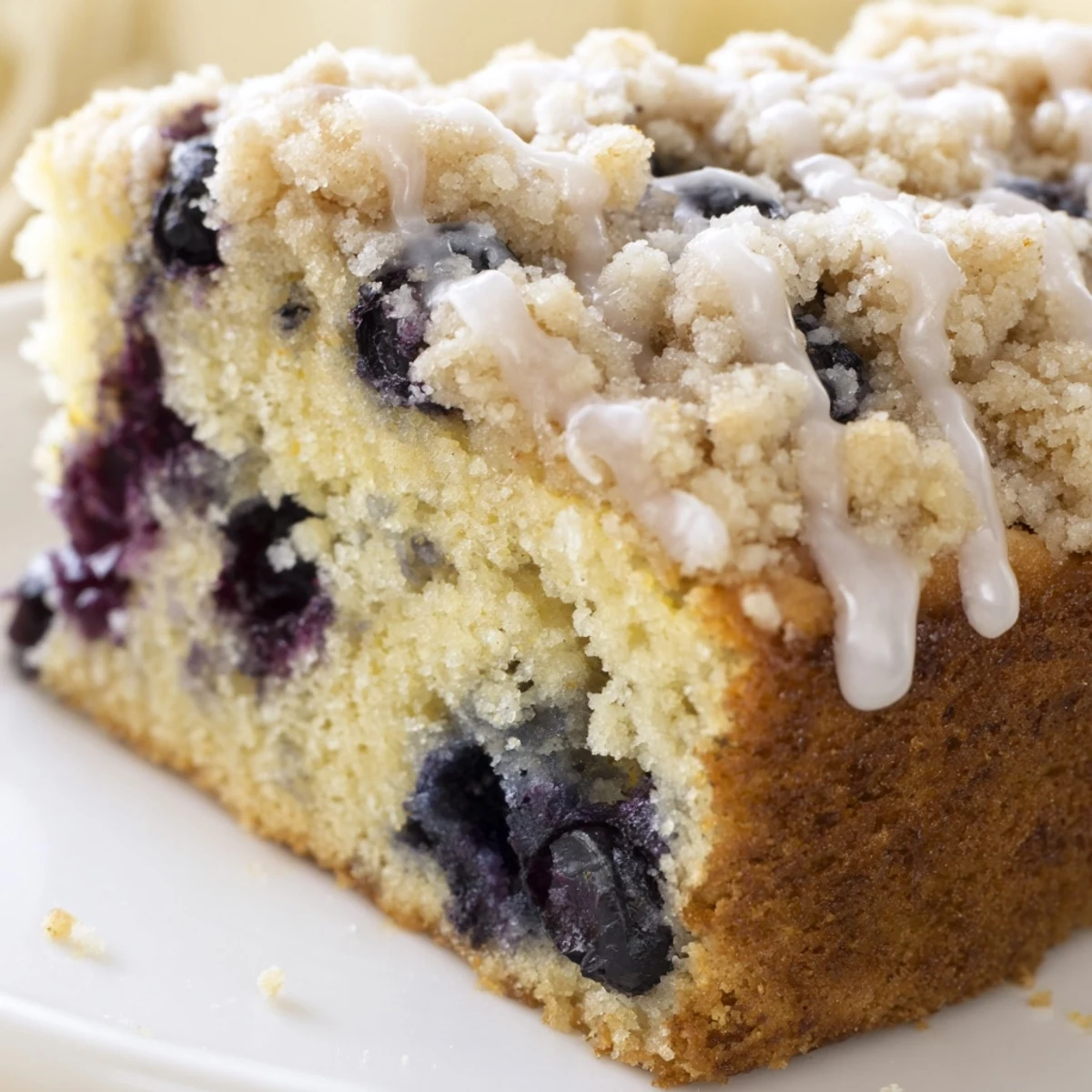 Golden-brown Lemon Blueberry Coffee Cake on a white plate, drizzled with a translucent lemon glaze and served with coffee.