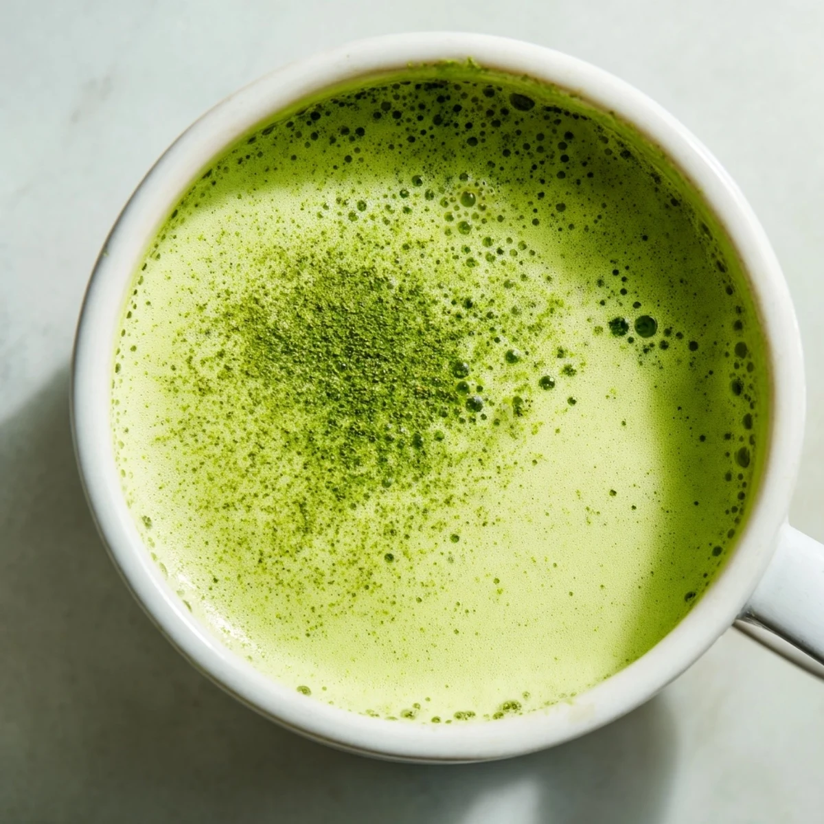 Green Matcha Latte with Almond Milk in a clear glass mug, dusted with vibrant green powder next to a bamboo whisk.