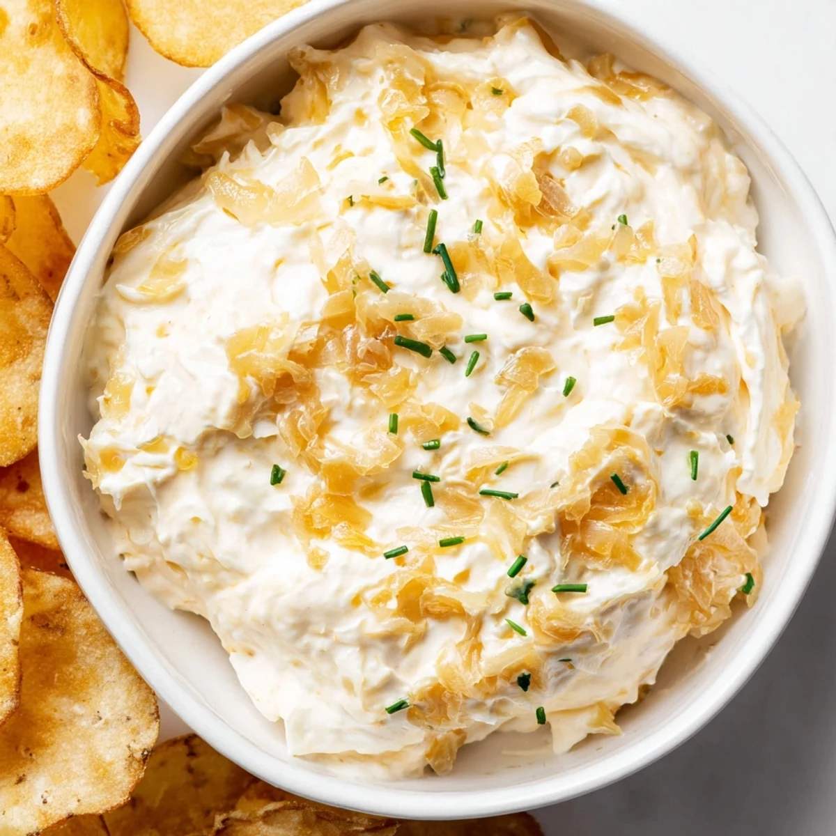 Golden brown, freshly caramelized onions folded into a rich dip with chives, surrounded by crunchy potato chips ready for dipping.