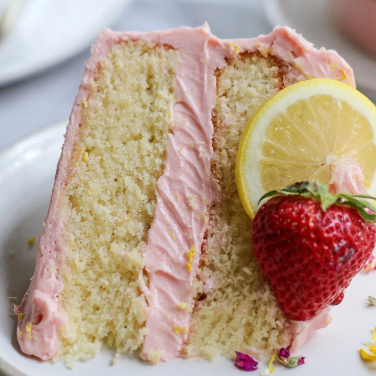 A slice of strawberry lemonade cake topped with pink frosting and fresh berry garnish on a rustic plate