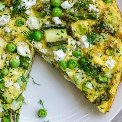 A freshly baked slice of Spring Vegetable Frittata with Herbs on a white plate, highlighting the bright green vegetables and melted Parmesan for a perfect spring brunch.  